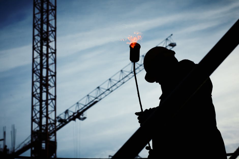 Silhouette of a construction worker using a blowtorch at a building site against a crane-filled skyline