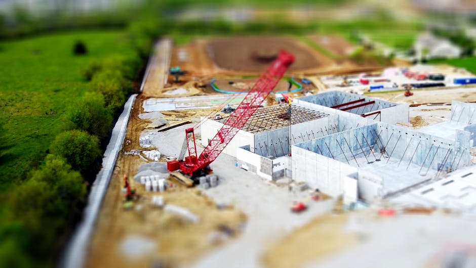 Aerial view of a construction site with a red crane amidst green fields and concrete buildings